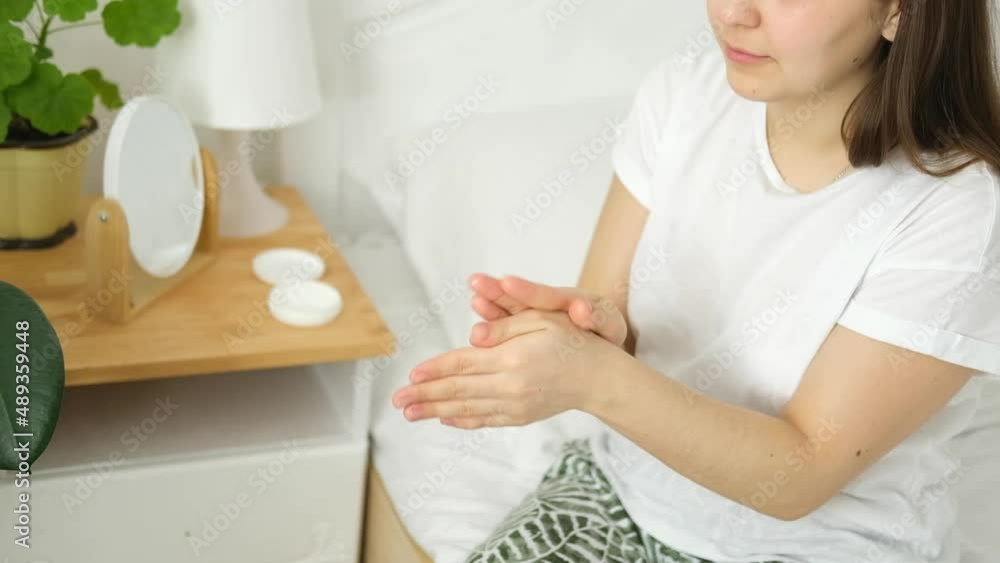 Young caucasian woman, brunette girl, is sitting on bed and smearing her hands with moisturizer. Home cosmetic spa care, hand massage. Young caucasian woman, brunette girl, is sitting on bed and smearing her hands with moisturizer. Home cosmetic spa care, hand massage.