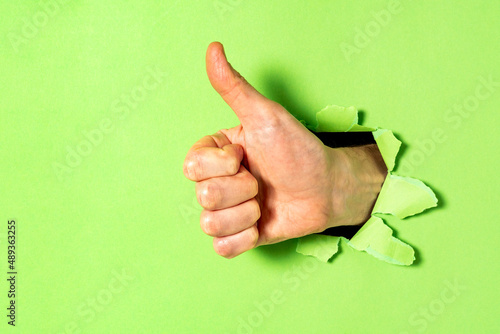 Hand with thumb up gesture through  hole in green paper wall photo