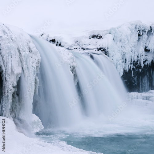 Goðafoss waterfall in winter. Ice formations around falls and snow on ground. Overcast sky. North Iceland.