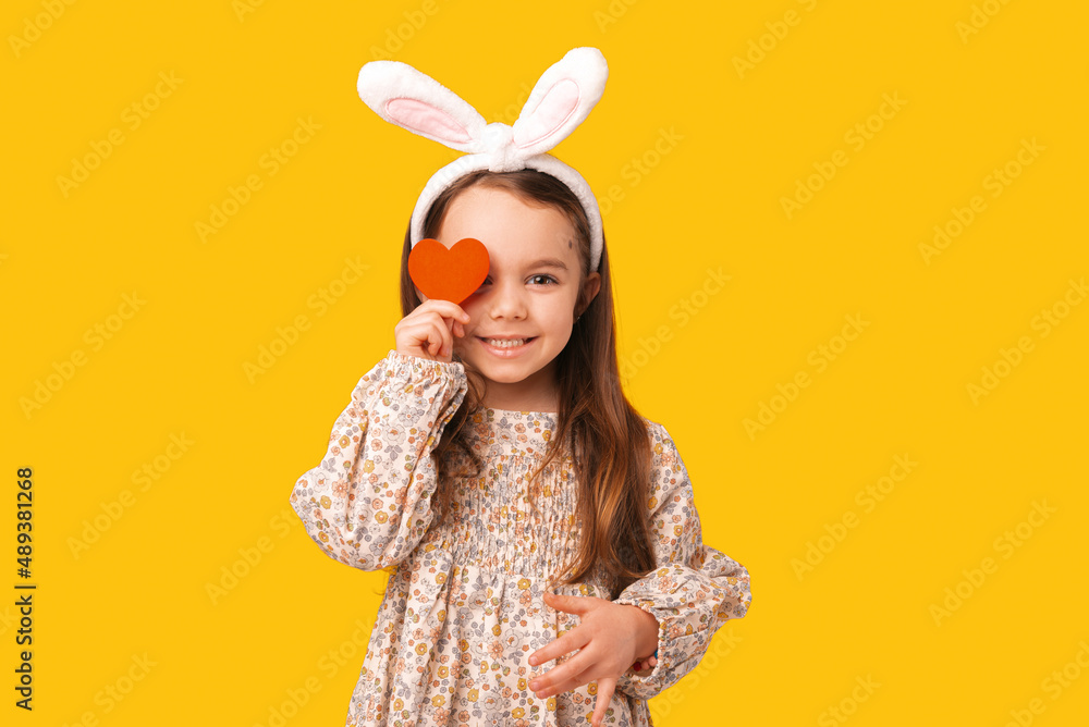 Cheerful little girl with bunny ears covers one eye with a red heart.
