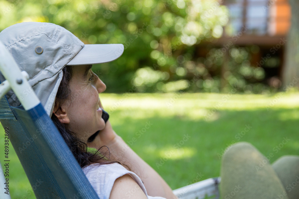 Close up of hispanic young woman wearing a cap talking with on mobile phone and doing home office while sitting in a lounge chair at the backyard in the springtime. Copy space.