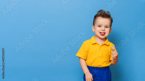 Baby boy with lollipop stands on a blue wall background. Prevention of childhood caries. Copy space