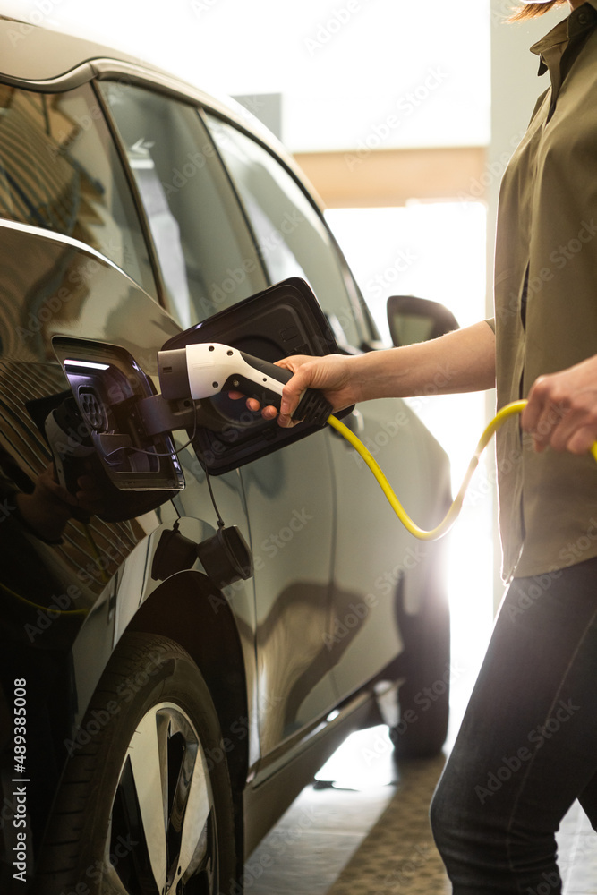Woman plugs a charger to electric black car socket at home garage. Woman plugs a charger to electric black car socket at home garage.