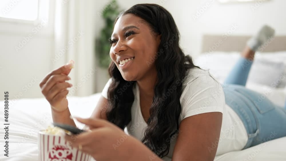 Happy Afro-American woman relaxing, eating popcorn while watching movie on bed Happy Afro-American woman relaxing, eating popcorn while watching movie on bed
