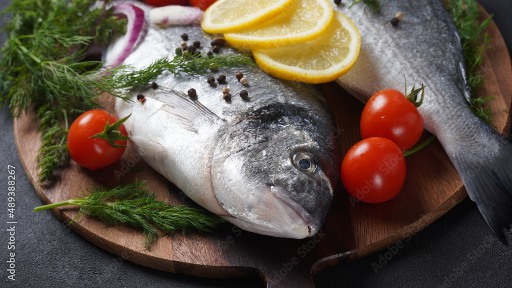 Raw fresh uncooked dorado or sea bream fish with lemon, herbs, cherry tomatoes and spices on black background Raw fresh uncooked dorado or sea bream fish with lemon, herbs, cherry tomatoes and spices on black background