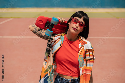 Mujer caucásica con pelo corto, tatuajes en el cuerpo y gafas de sol en una pista deportiva con un skate 