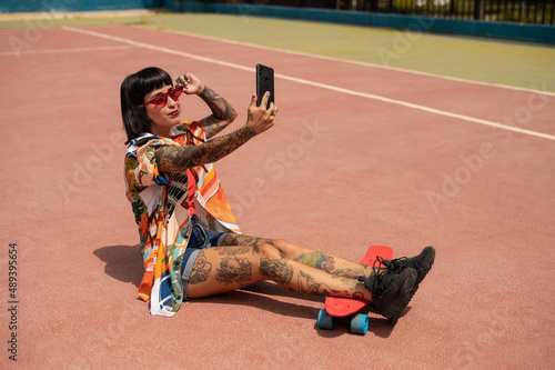 Caucasian woman with tattoos and short hair on a sports court with a skateboard using her phone's camera