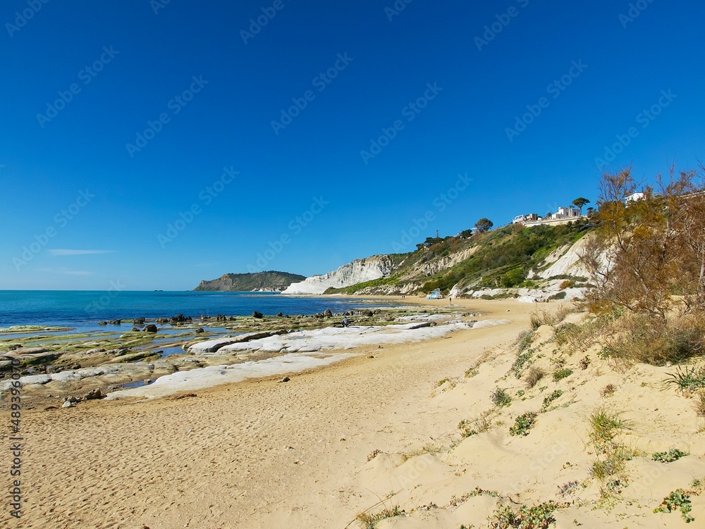 a coastline view with sand beach, rocks, and a green hill in Realmonte, Agrigento against the blue sky on a clear sunny day, Sicily a coastline view with sand beach, rocks, and a green hill in Realmonte, Agrigento against the blue sky on a clear sunny day, Sicily