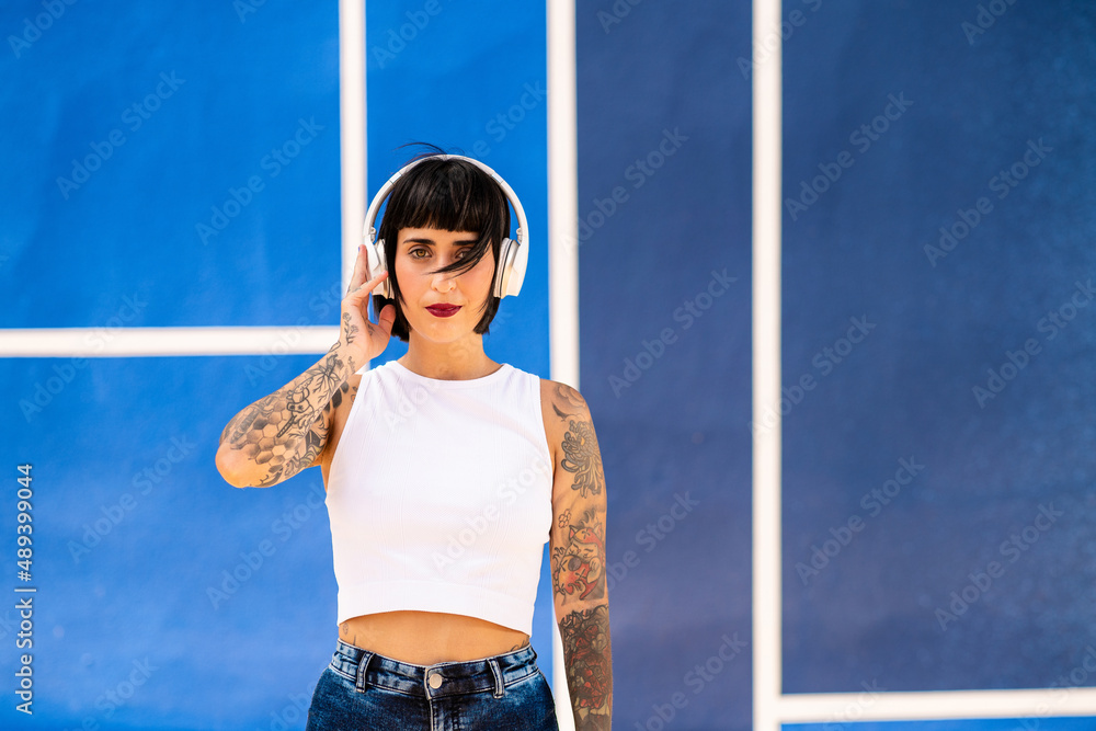 Caucasian woman with tattoos on her arms and short hair listening to music with headphones on a sports court