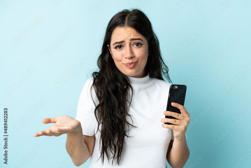 Young woman using mobile phone isolated on blue background making doubts gesture while lifting the shoulders Young woman using mobile phone isolated on blue background making doubts gesture while lifting the shoulders