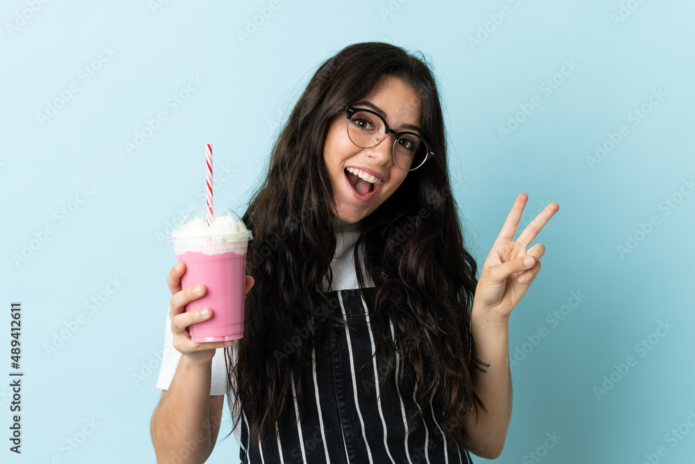 Young woman with strawberry milkshake isolated on blue background smiling and showing victory sign Young woman with strawberry milkshake isolated on blue background smiling and showing victory sign