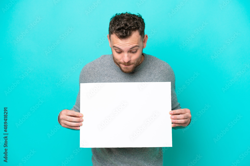 Young Brazilian man holding an empty placard isolated on blue background holding an empty placard and looking it Young Brazilian man holding an empty placard isolated on blue background holding an empty placard and looking it