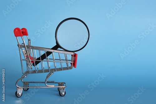 Magnifying glass inside shopping cart. Blue background. Product search and frugal shopping concept.