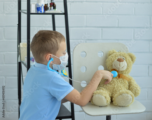 A boy in a blue shirt is playing doctor  listening to the heart of a toy bear with a phonendoscope.