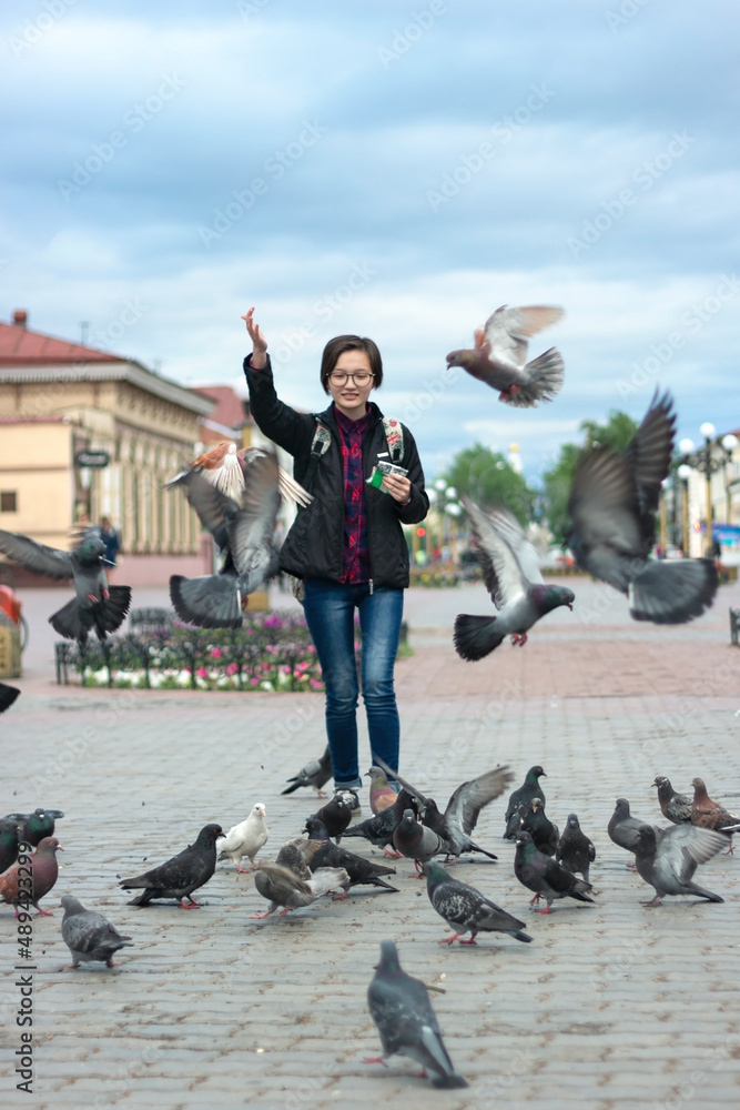Obraz premium Young woman teenager feeding pigeons with seeds on wide pedestrian city street at cloudy day, urban bird catches food on fly, vertical image