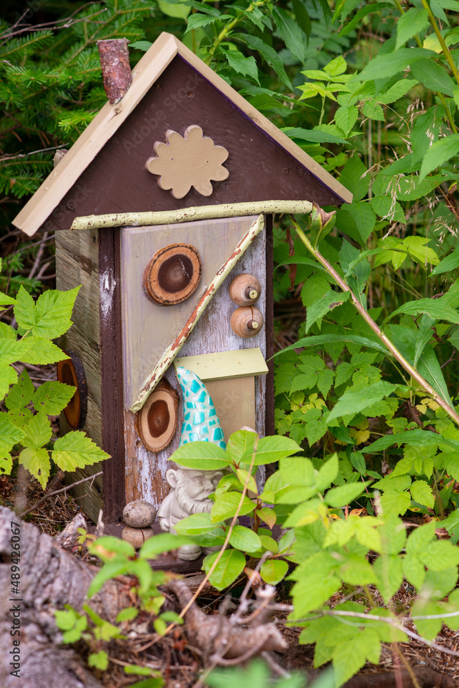 A small ceramic garden gnome with a blue hat stands next to a tiny colorful fantasy house with windows, a door and a slanted roof. The magical wooden building is among green garden shrubs and plants. A small ceramic garden gnome with a blue hat stands next to a tiny colorful fantasy house with windows, a door and a slanted roof. The magical wooden building is among green garden shrubs and plants.