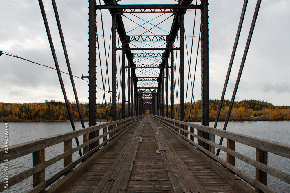 Steel trestle bridge with wooden deck over a large river in Newfoundland.  The bridge is for foot traffic and ATV usage. The sky is clear blue and land and houses can be seen in the background