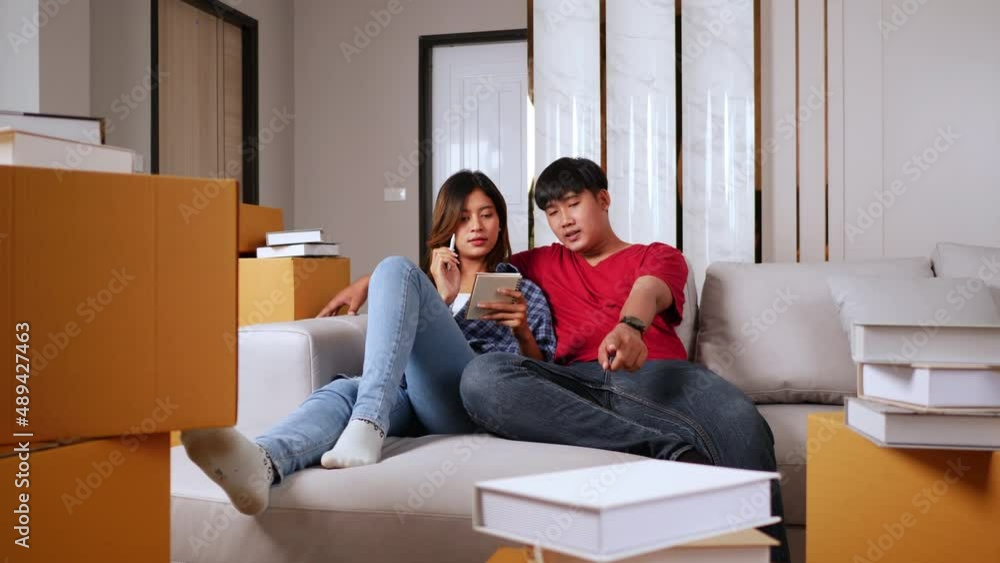 Slow motion shot, Young couple sitting on sofa bed and checking list while count the boxs after relocation in their new home, new house concept Slow motion shot, Young couple sitting on sofa bed and checking list while count the boxs after relocation in their new home, new house concept