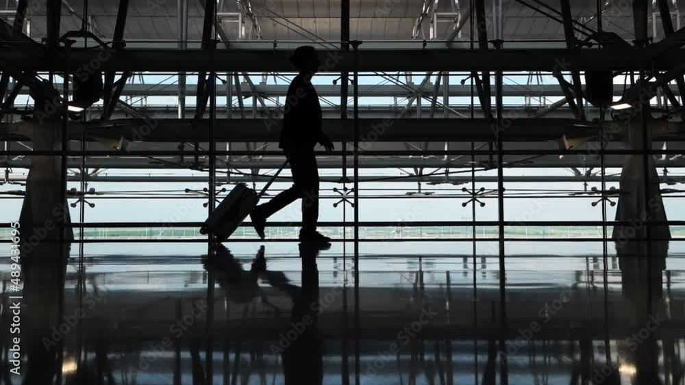 Passenger man go to boarding gate with small wheeled case. Silhouetted shot, slow motion, full length view against window. Unhurried guy walks to the departure area through modern terminal building Passenger man go to boarding gate with small wheeled case. Silhouetted shot, slow motion, full length view against window. Unhurried guy walks to the departure area through modern terminal building