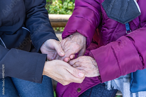 A mature woman holds her elderly mother's hands as they sit on a bench