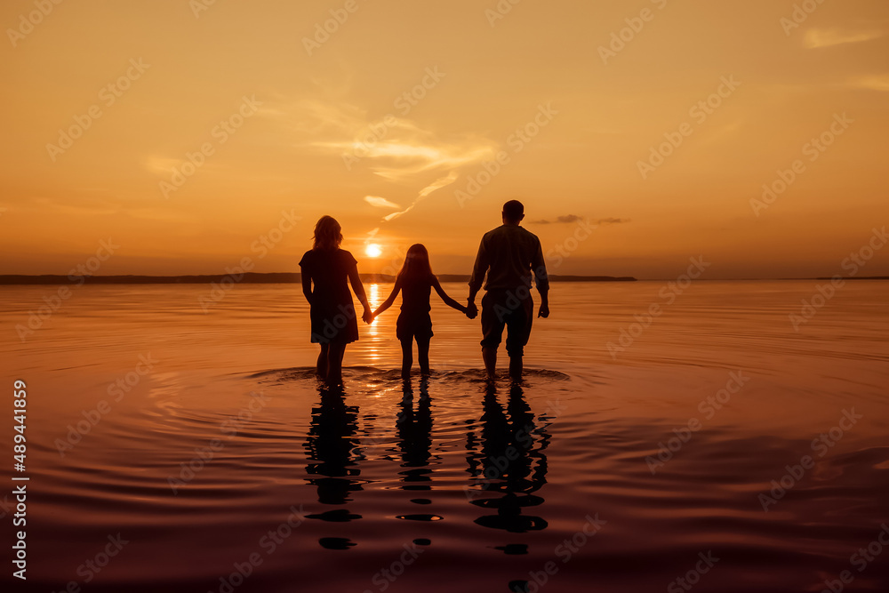Full body silhouette of an unrecognizable family holding hands and walking with their backs to the camera on the water of a river, during sunset, copy space, toned image