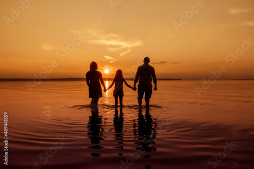 Full body silhouette of an unrecognizable family holding hands and walking with their backs to the camera on the water of a river  during sunset  copy space  toned image