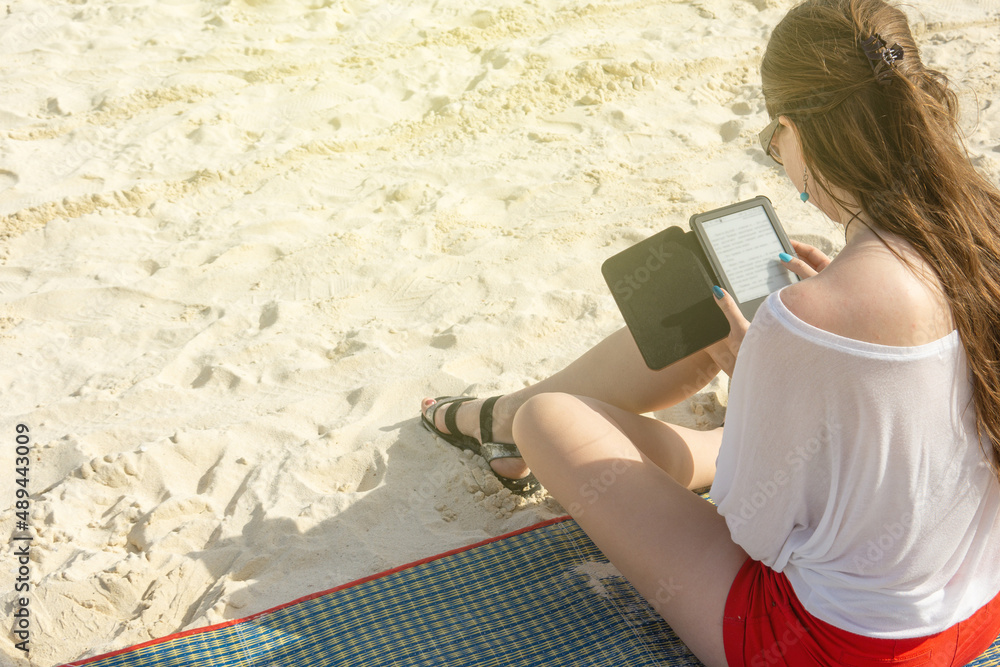 A girl reads an e-book sitting on a sandy beach. A girl in sunglasses reads on the sand, view from the back. A girl reads an e-book sitting on a sandy beach. A girl in sunglasses reads on the sand, view from the back.