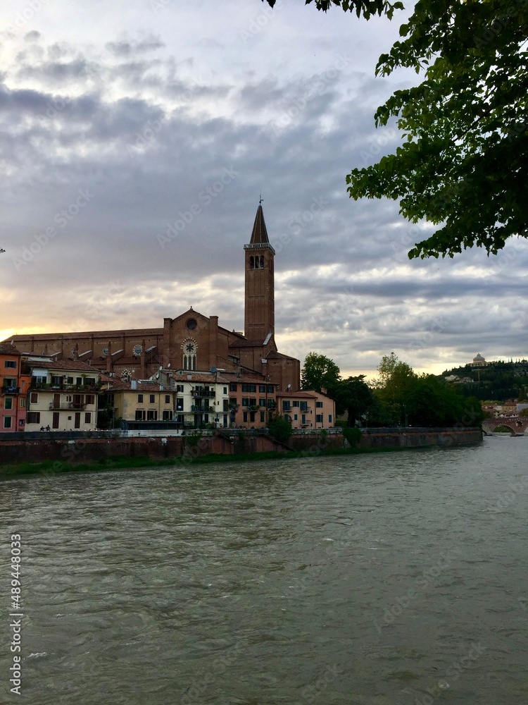 Verona, Italy. Town view from Nuovo del Popolo bridge over Adige river. Sunset, Bell tower of Santa Anastasia church. Cloudy sky. Green trees.