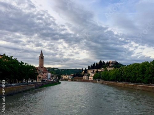 Verona, Italy. Town view from Nuovo del Popolo bridge over Adige river. Sunset, Bell tower of Santa Anastasia church. Cloudy sky. Green trees © Alla