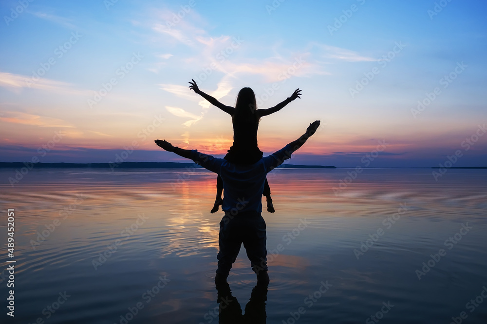 Full length silhouettes of an unrecognizable standing man and a little girl, sitting on his shoulders and raising their hands against the evening sky in the water of the river during sunset in summer