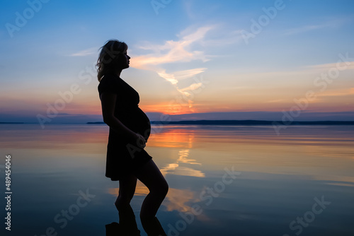 Full length silhouette unrecognized female expecting a baby  who stands in the water of the lake and touch her belly against evening sky background at sunset. Pregnancy  copy space 