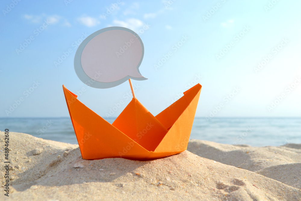 Orange Paper Boat on Sandy Seashore Close-up. Small Paper Boat on Background of Blue Sea on Sunny Summer day. Stick with empty white blank speech bubble in Boat. Concept Travel, Tourism, Vacation Rest Orange Paper Boat on Sandy Seashore Close-up. Small Paper Boat on Background of Blue Sea on Sunny Summer day. Stick with empty white blank speech bubble in Boat. Concept Travel, Tourism, Vacation Rest