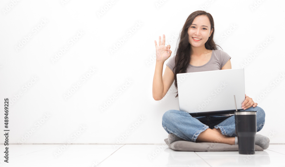 Beautiful Asian woman wearing grey casual shirt using laptop on white background and copy space. Cute Asian woman sitting on the floor room and okay sign relaxing for work from home concept Beautiful Asian woman wearing grey casual shirt using laptop on white background and copy space. Cute Asian woman sitting on the floor room and okay sign relaxing for work from home concept