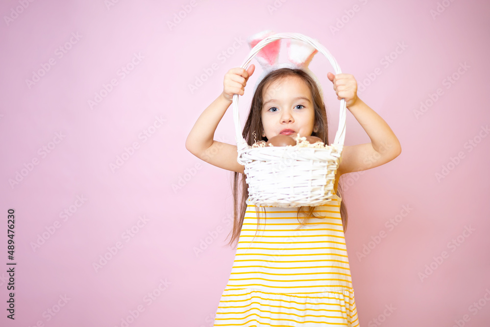 Happy Easter. A cute funny girl with bunny ears holds a basket of chocolate eggs in her hands. Pink background. Happy Easter. A cute funny girl with bunny ears holds a basket of chocolate eggs in her hands. Pink background.
