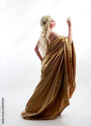 Full length portrait of pretty female model wearing grecian goddess toga gown, posing with elegant gestural movements on a studio background.