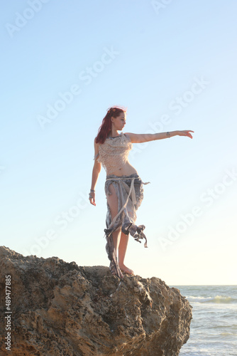  portrait of pretty female ship wrecked model wearing torn dress. posing on the rocky Ocean shoreline at sunset,