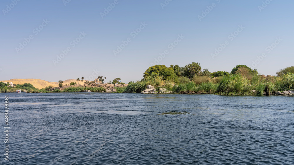 Lush green vegetation, picturesque boulders, sand dunes are visible on the banks of the Nile. Ripples on the blue water. Clear azure sky. A sunny day. Egypt.