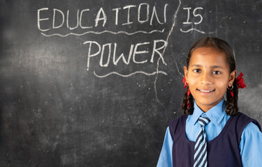 Smiling young girl kid with school uniform looking camera after writing education is power on black board at class room - concept of development, growth and wisdom