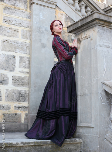 Full length portrait of red-haired woman wearing a historical victorian gown costume, walking around beautiful location with  Gothic stone architecture. © faestock