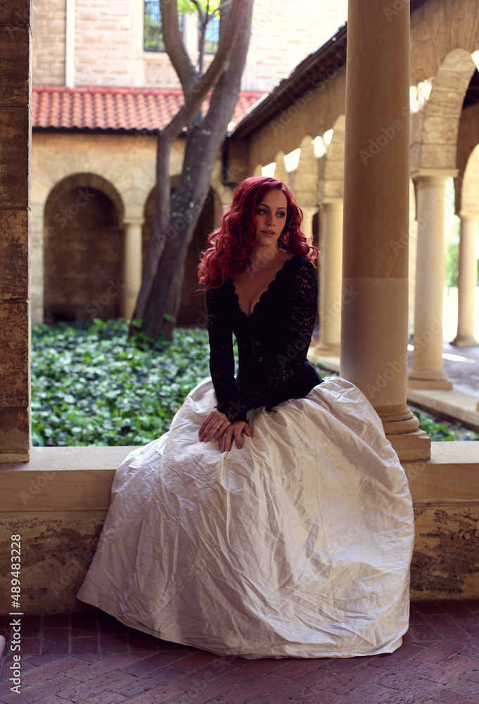 Full length portrait of red-haired woman wearing a  beautiful gothic gown costume, walking around  location with  romantic castle stone architecture background.