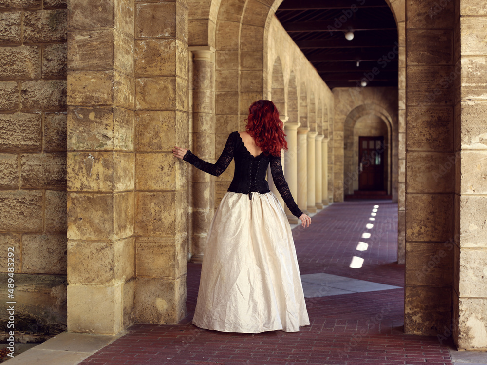 Full length portrait of red-haired woman wearing a beautiful gothic gown costume, walking around location with romantic castle stone architecture background. Full length portrait of red-haired woman wearing a beautiful gothic gown costume, walking around location with romantic castle stone architecture background.