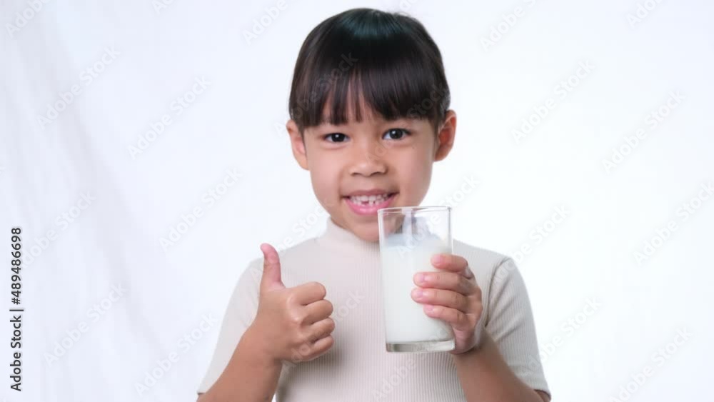 Cute Asian little girl holding a glass of milk and showing thumbs up on white background in studio. Healthy nutrition for small children. Cute Asian little girl holding a glass of milk and showing thumbs up on white background in studio. Healthy nutrition for small children.