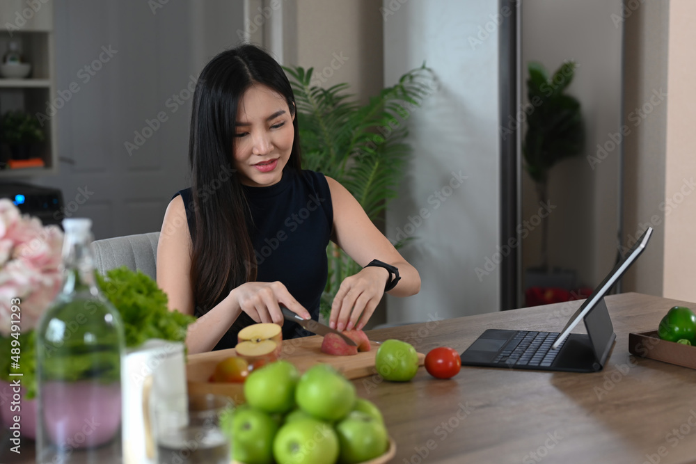 Beautiful woman cutting apple on wooden board and reading recipe on digital tablet.