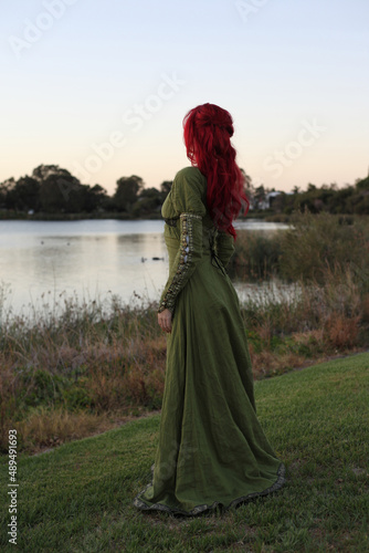 Full length portrait of red haired woman wearing a beautiful green medieval fantasy gown. Posing with gestural hands on a enchanted forest background.