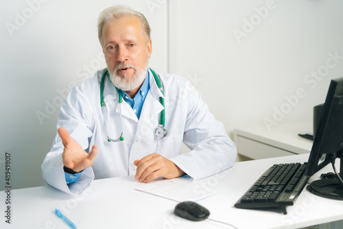 Portrait of confident mature adult male doctor in white uniform coat with stethoscope speaking, looking at camera, gesturing sitting at desk with computer in medical office, consulting patient.