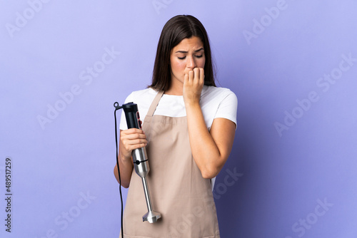 Brazilian woman using hand blender isolated on purple background having doubts