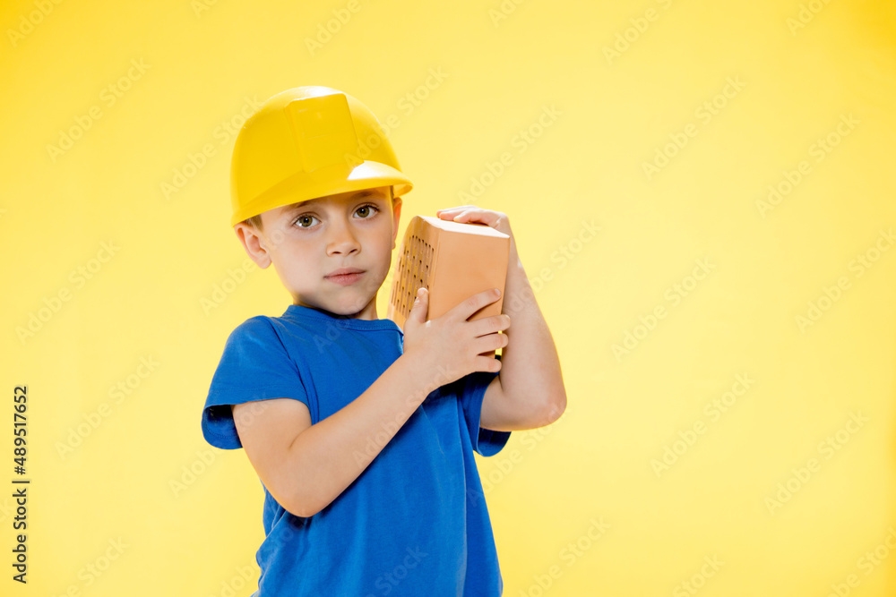 Boy in a construction helmet holds a brick in his hands on yellow background Boy in a construction helmet holds a brick in his hands on yellow background