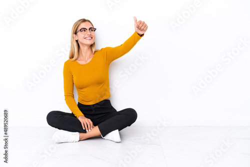 Blonde Uruguayan girl sitting on the floor giving a thumbs up gesture