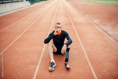 A tired runner with artificial leg sitting on running track and drinking refreshment.