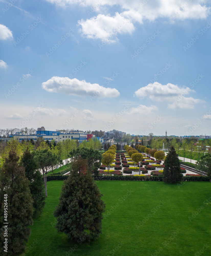 An elegant city park under a blue sky with white clouds. Top view of a beautiful park in early spring. Urban landscape. Green lawn near the alley with trees
